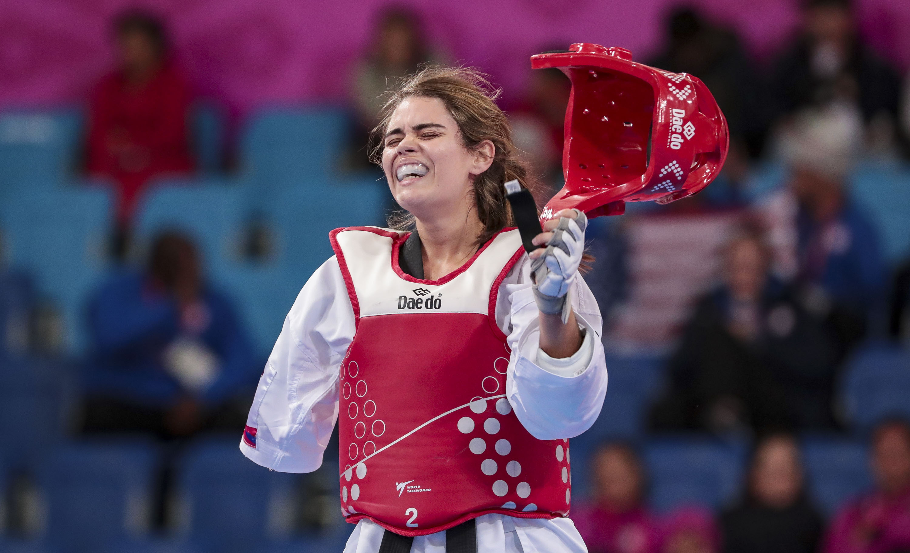 Na foto horizontal, uma jovem de pele clara e cabelos castanhos e com expressão de cansaço veste um uniforme de taekwondo branco e vermelho. Ela não tem a mão direita, por isso a manga desse lado está solta. Na mão esquerda, ela segura um capacete vermelho para cima que, aparentemente, acabou de tirá-lo da cabeça. Ao fundo, em imagem desfocada, público assiste à luta