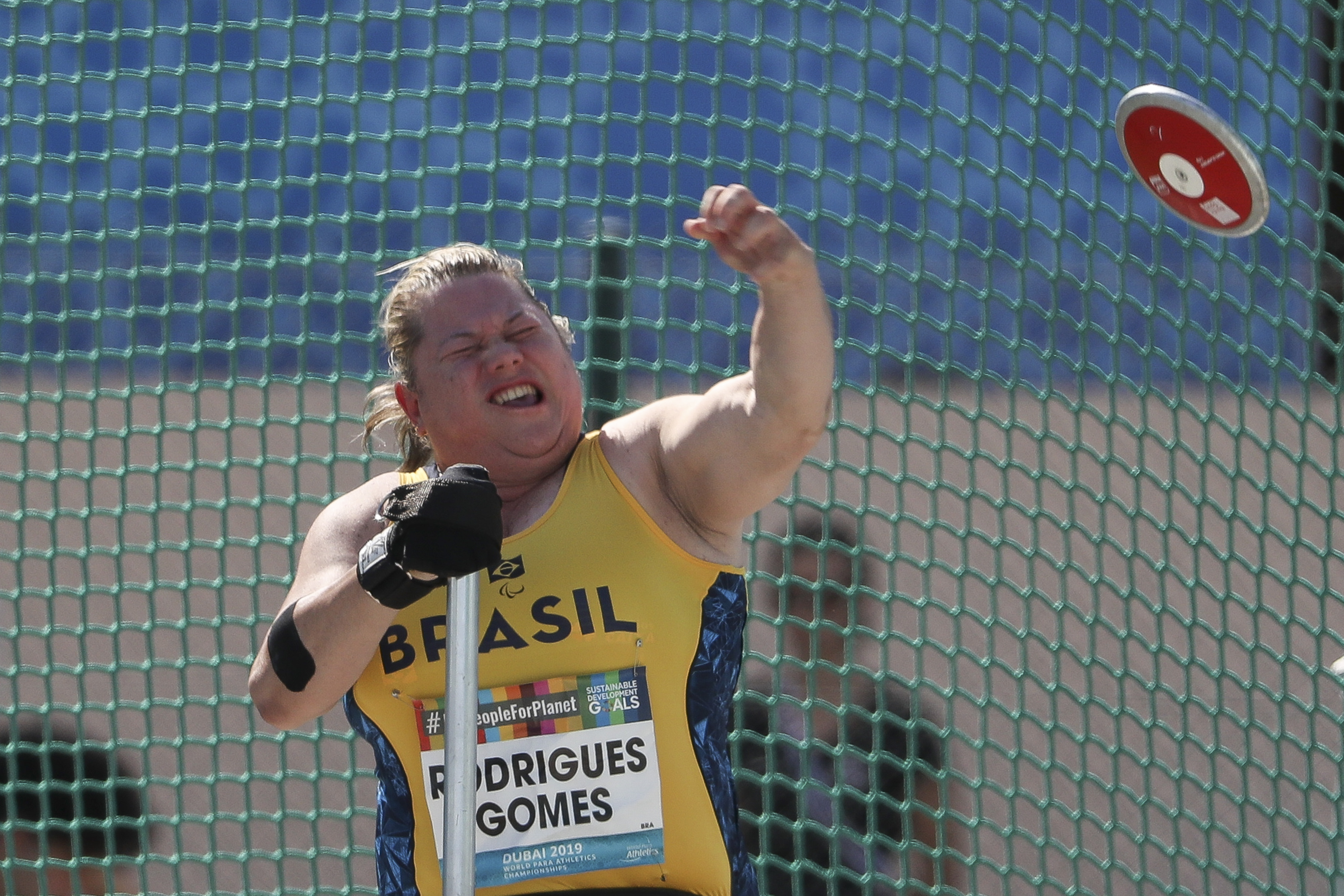 Na foto horizontal, uma atleta de meia idade loira, de pele clara e olhos azuis veste uniforme amarelo do Brasil. Ela faz uma careta de esforço com o braço esquerdo erguido e o braço direito apoiado em um taco cinza. No canto superior direito há um disco vermelho no ar