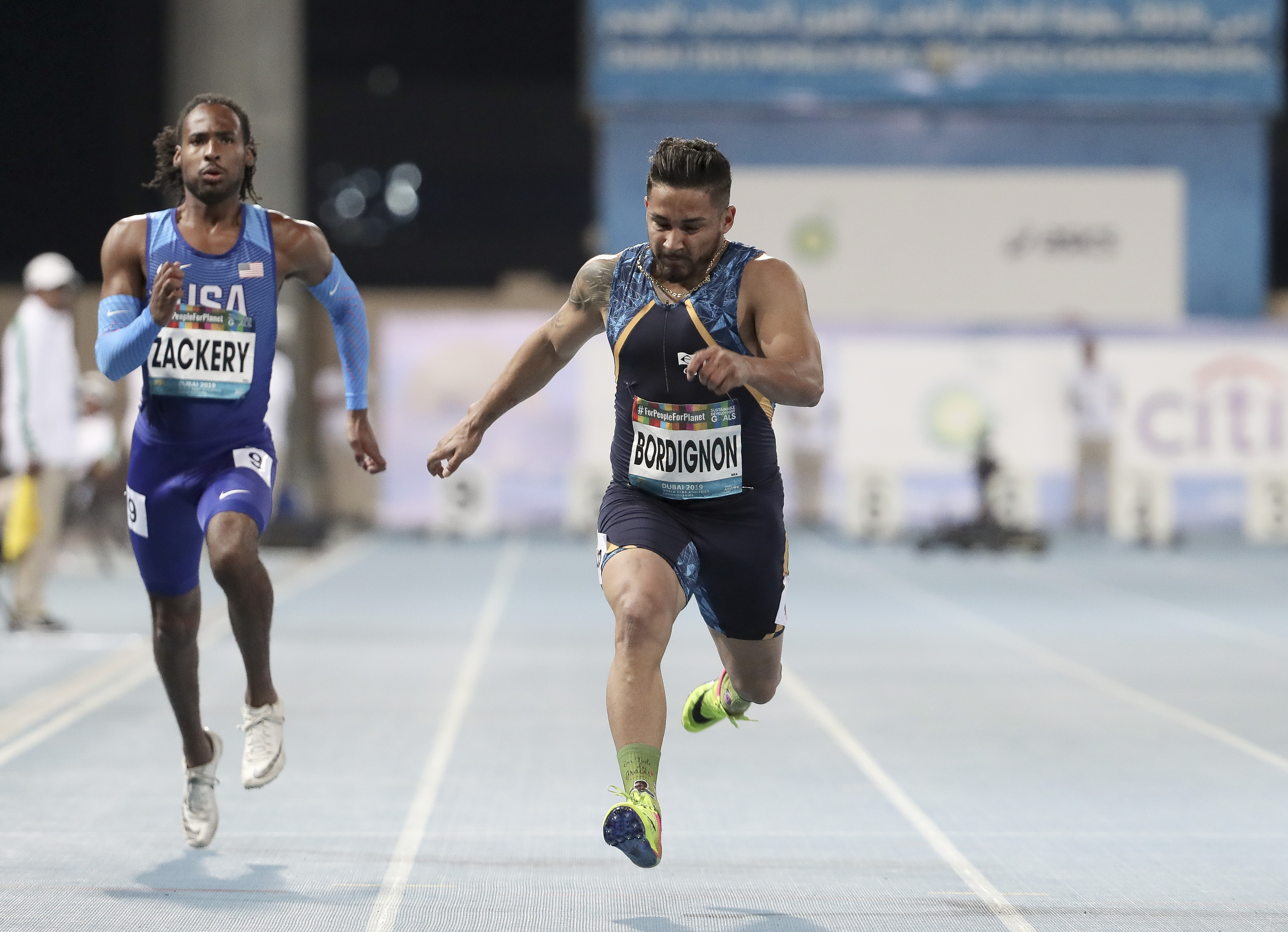  Na foto horizontal, ao centro, um atleta brasileiro de pele morena e cabelos curtos e barba corre vestindo um uniforme azul escuro e tênis amarelo. Ao seu lado esquerdo, um atleta norte-americano de pele negra corre vestindo um uniforme azul claro e tênis branco. Ambos correm um uma pista de atletismo azul e listas brancas.