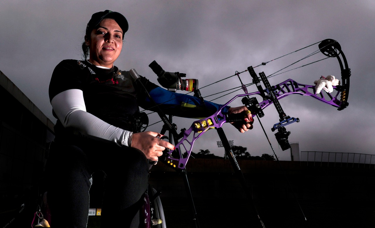 Na foto horizontal, uma mulher de meia idade está sentada em uma cadeira de rodas. Ela segura com as duas mãos o equipamento de arco e flecha. A atleta está no campo aberto do CT Paralímpico e com céu escuro e nublado ao fundo